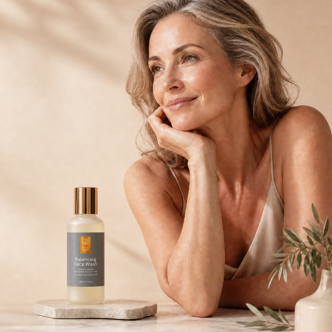 A woman in a beige satin top rests her head on her hand beside a bottle of Balancing Face Wash on a marble tray. The bottle has a gold cap and a dark gray label with a gold shield logo.